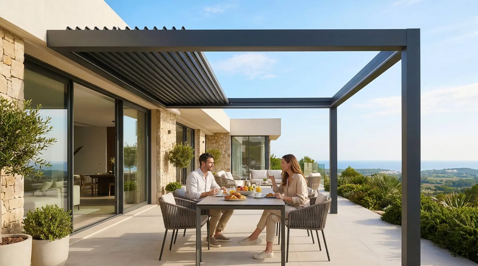 Luxa 800 aufschiebbares Lamellendach — Paar beim Frühstück auf mediterraner Terrasse mit gekippten Lamellen und Meerblick